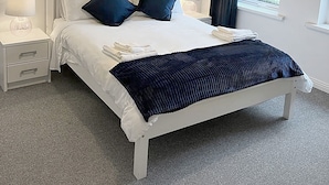 A neatly made bed with white bedding and two dark blue pillows, a white bedside table with a lamp, and a window with a view of greenery. - Burren Seaside Lodge (Ballyvaughan)