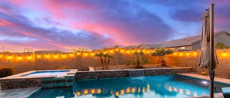 A swimming pool with a waterfall feature, surrounded by a stone wall and palm trees, under a twilight sky.