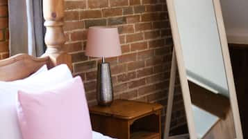 A bed with white and pink pillows, a wooden nightstand with a lamp, and a large mirror leaning against a brick wall.
