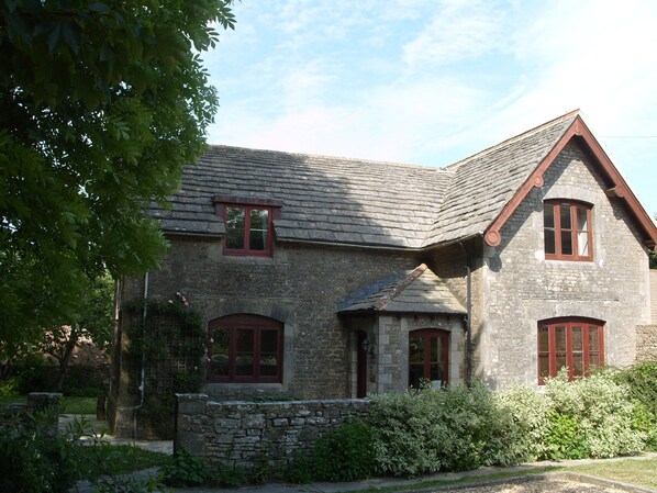 A stone building with a steeply pitched roof, multiple windows with red frames, and a stone wall in front. - Kingston Country Courtyard (Wareham)