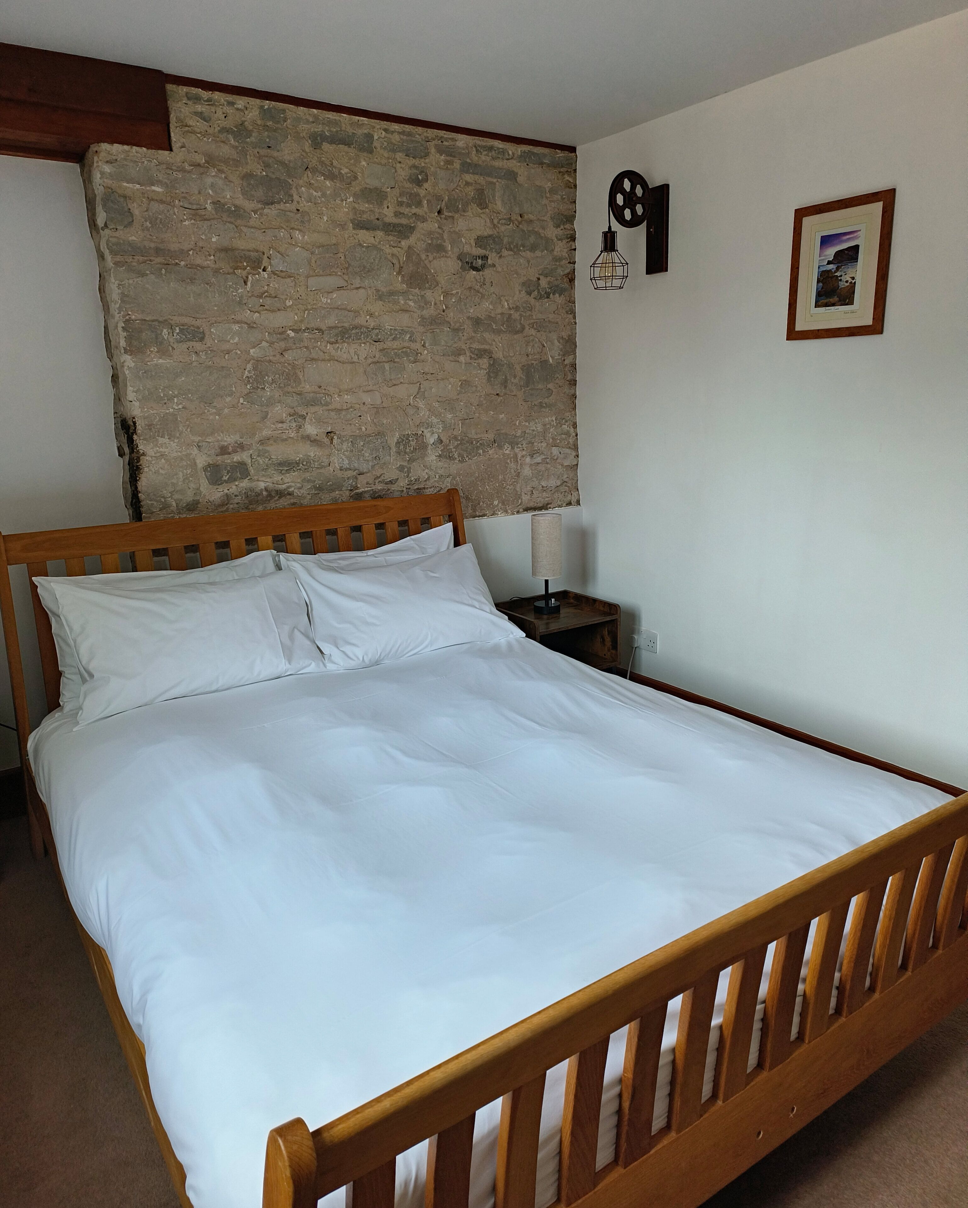 Un lit simple avec du linge de lit blanc, une tête de lit en bois, une table de chevet avec une lampe et un mur en pierre.