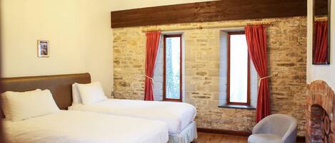 A room with two beds, a stone wall, wooden beams, a chandelier, and a brick fireplace.