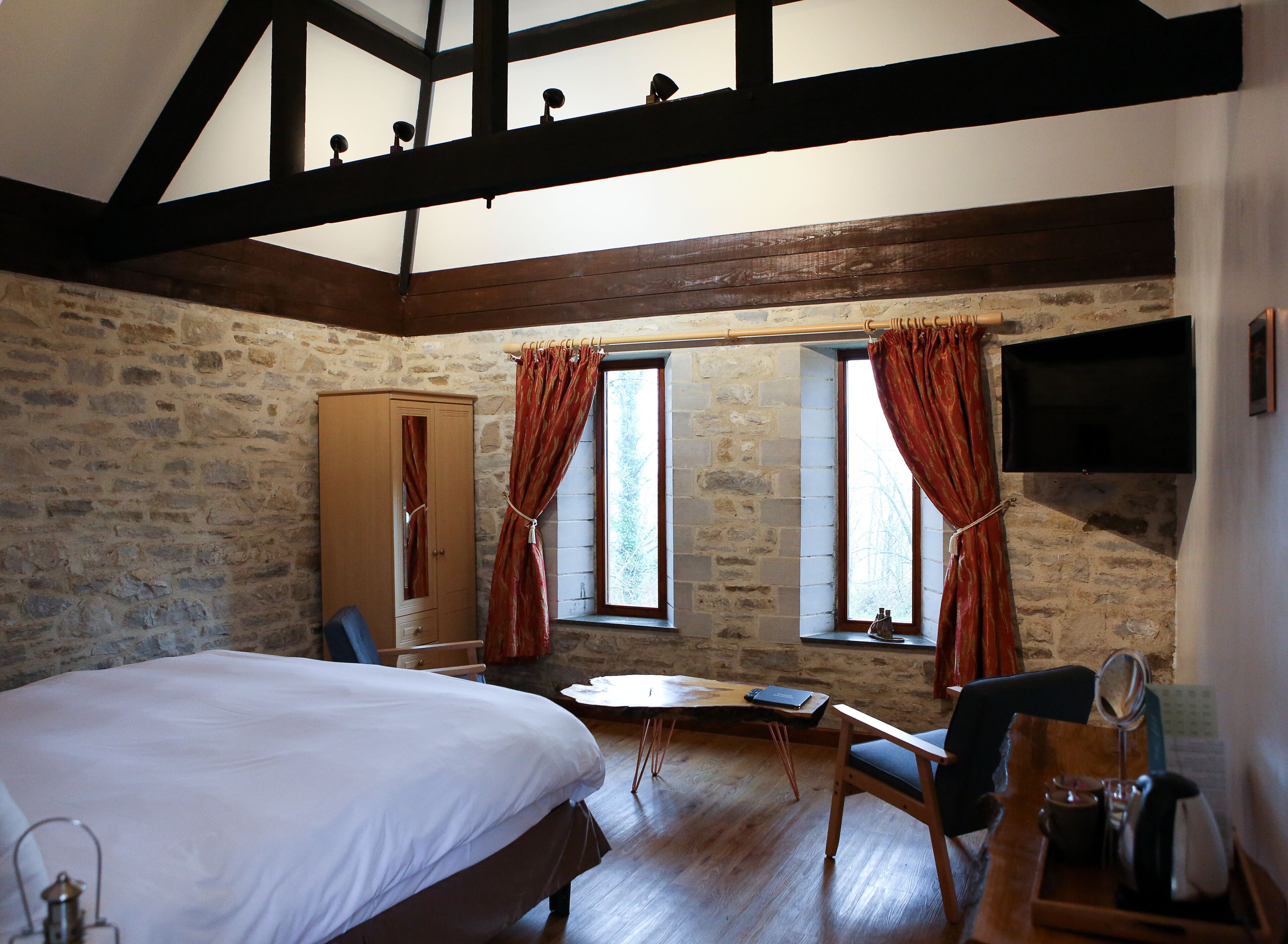 A bedroom with a large bed, wooden beams, stone walls, a wooden desk, and a television mounted on the wall.