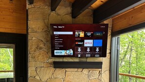 Un téléviseur TCL Roku fixé au mur en pierre, au-dessus d’une cheminée, avec un plafond en bois et une vue sur les arbres à travers une porte vitrée.