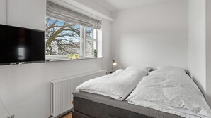 Une chambre à coucher avec un lit, un téléviseur fixé au mur, une fenêtre donnant sur des arbres et un radiateur.