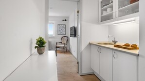 Une cuisine étroite avec des meubles blancs, un plan de travail en bois et un évier. On trouve une chaise et une plante en pot dans le couloir.