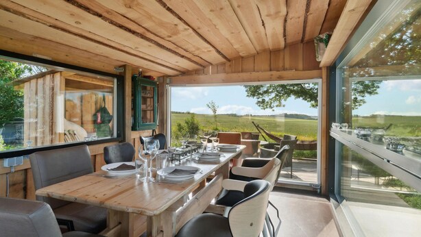Un coin repas en bois avec une table dressée pour quatre personnes, de grandes fenêtres offrant une vue sur un paysage verdoyant et une décoration intérieure rustique.
