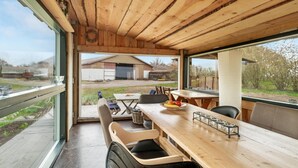 Une terrasse en bois avec une table et des chaises, une vue sur une grange et des arbres, et une porte vitrée donnant sur l’extérieur.