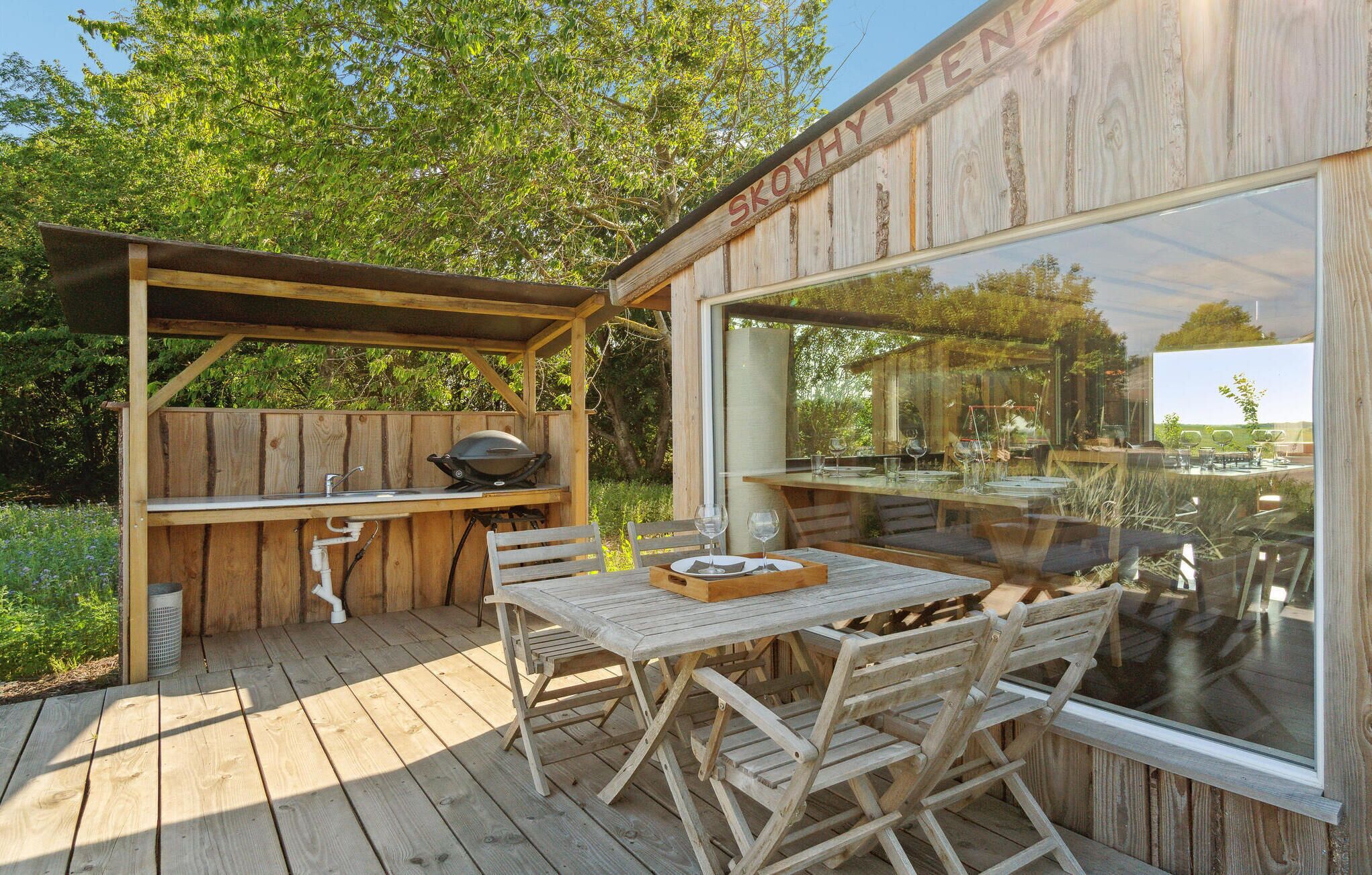 Een houten terras buiten met een barbecuehoek, een eettafel met stoelen en een groot raam met uitzicht op de omgeving.