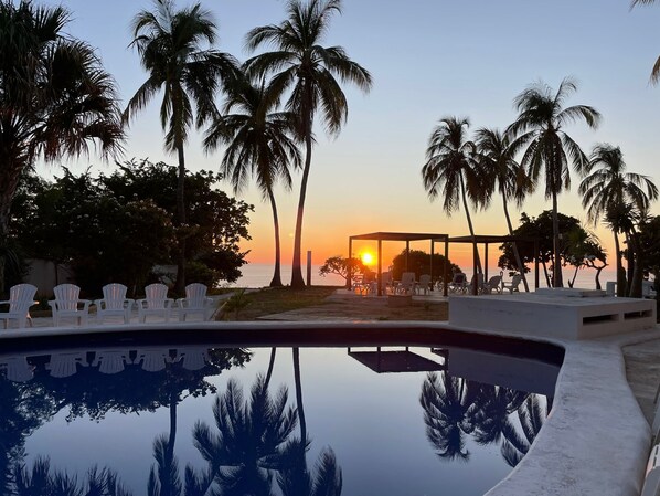 Vista a la piscina con palmeras, un atardecer y una zona de estar cubierta.