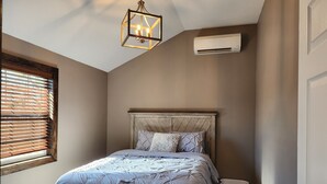 Une chambre avec une tête de lit en bois, un lit recouvert d’une couette à motifs, un luminaire suspendu et un panier à linge.