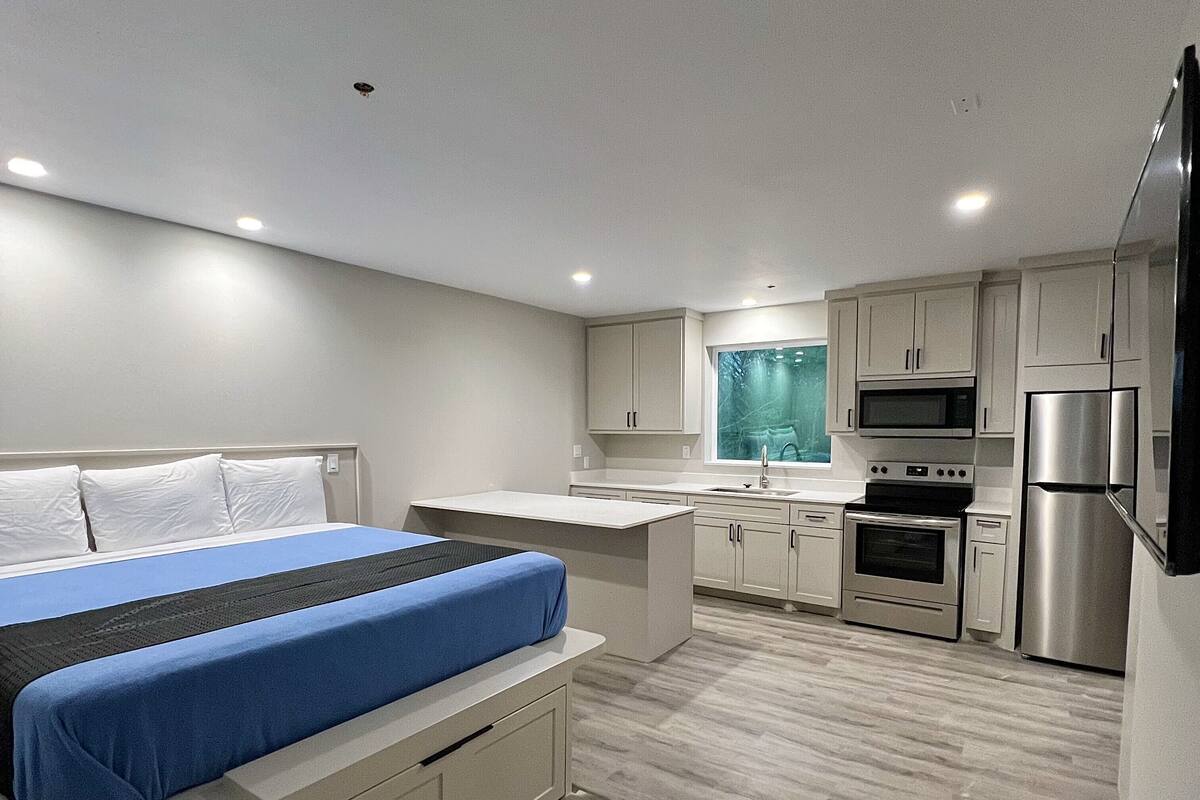 A modern bedroom with a bed, a small desk, and a kitchenette with a microwave, oven, and refrigerator.