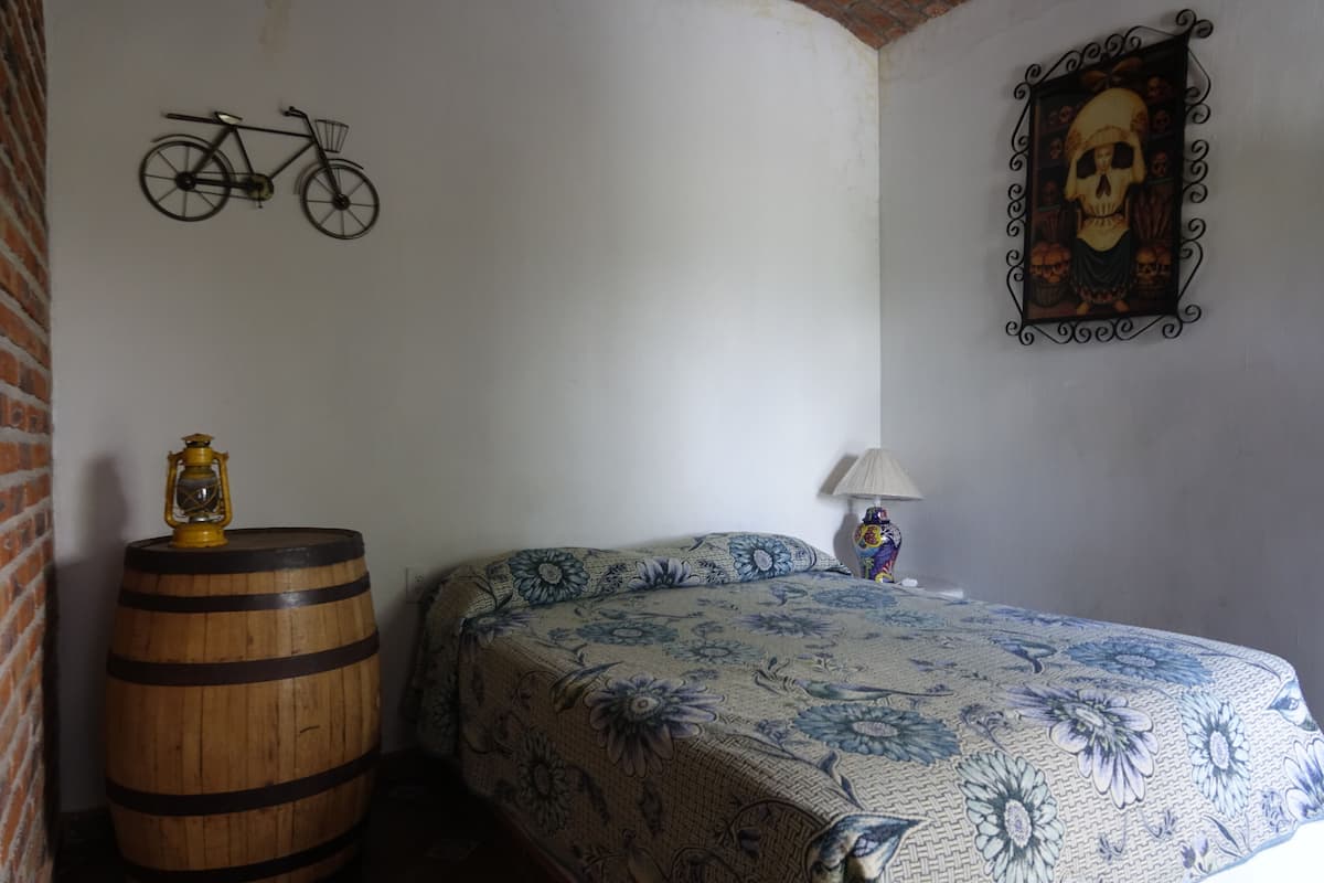 Un dormitorio con cubrecama floral, un barril de madera, una bicicleta colgada en la pared, una decoración de calavera y una lámpara.