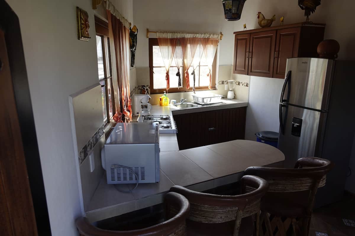 Cocina compacta con estufa a gas, lavamanos y refrigerador. Hay sillas de mimbre y una pequeña mesa de comedor.