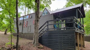 A cabin with a wooden deck, a covered porch, and a view of the surrounding forest. - Juniper: Sleep 5, Tiny Cabin, Pets, Near Park/Lake (Broken Bow)