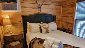 A wooden cabin room with a bed, a nightstand, a lamp, and a deer-themed pillow. - Juniper: Sleep 5, Tiny Cabin, Pets, Near Park/Lake (Broken Bow)