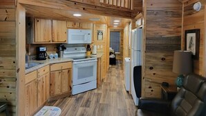 A cabin interior with wooden walls, a kitchen with a stove, microwave, and refrigerator, and a seating area with a chair. - Juniper: Sleep 5, Tiny Cabin, Pets, Near Park/Lake (Broken Bow)