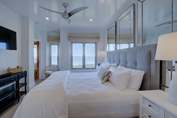 Ein geräumiges Schlafzimmer mit einem großen Bett, einem Deckenventilator, einem Fernseher und Blick auf den Strand durch die Fenster.