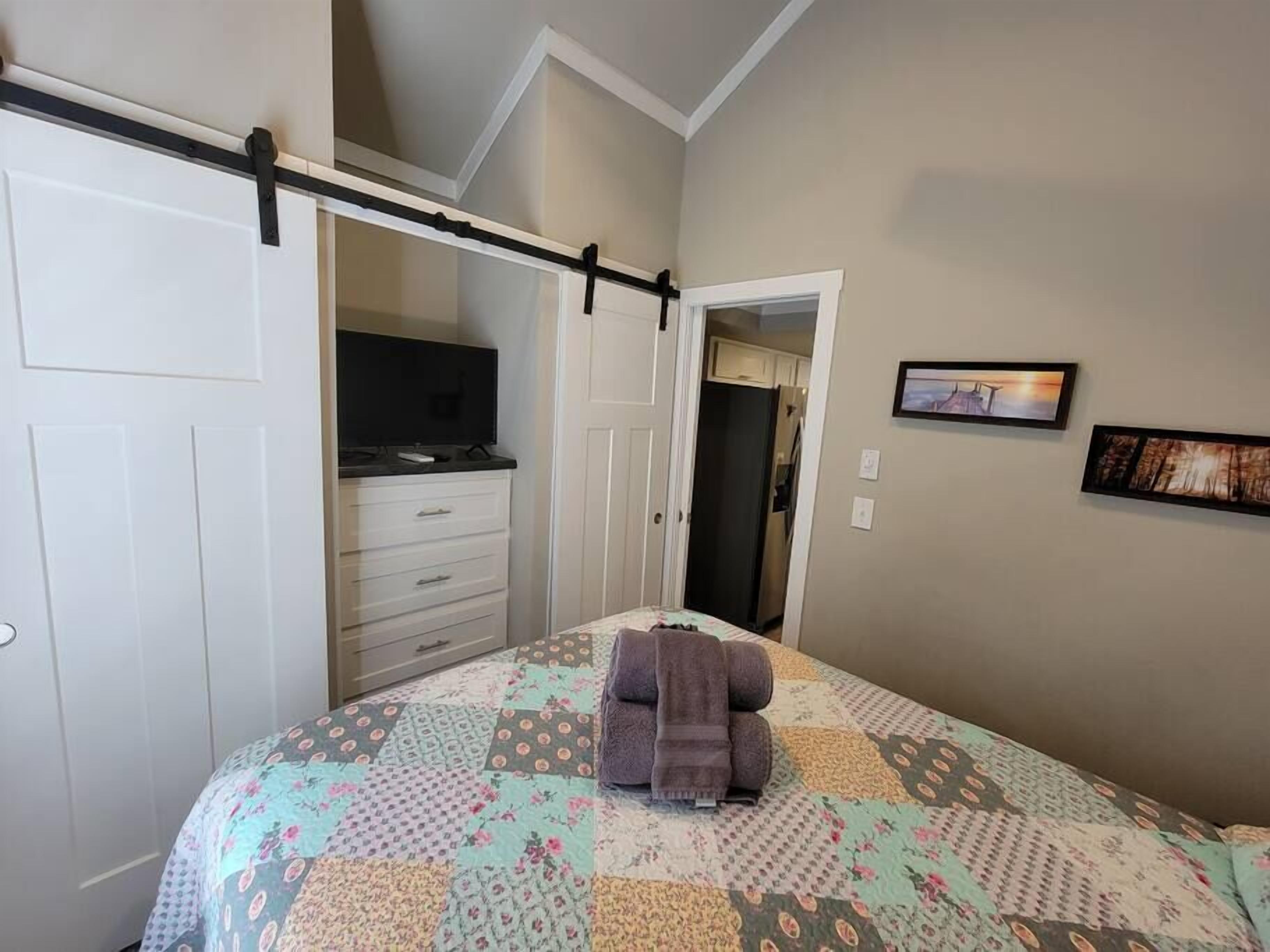 Ein Schlafzimmer mit einem Bett, einem Fernseher auf einer Kommode, einer Tür zu einem anderen Raum und einem gerahmten Bild an der Wand.