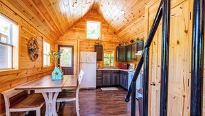 A wooden cabin interior with a dining table, chairs, a refrigerator, cabinets, and a staircase. - Acorn: Sleeps 4, Tiny Cabin, Pets, Near Park/Lake (Broken Bow)