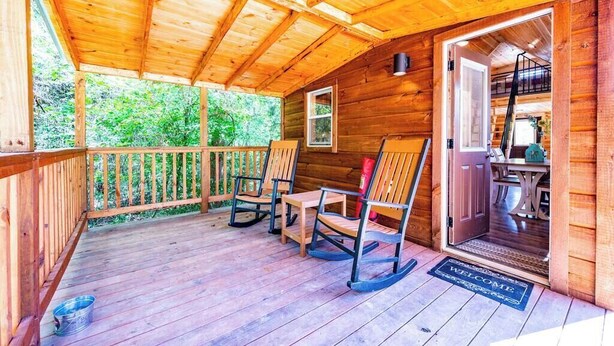 A wooden cabin with a porch, rocking chairs, and a door leading to an interior with a table and chairs. - Acorn: Sleeps 4, Tiny Cabin, Pets, Near Park/Lake (Broken Bow)