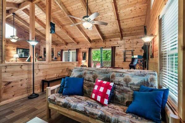 L’intérieur d’une cabane avec un plafond en bois, un canapé orné d’un motif représentant une forêt et un ventilateur de plafond.