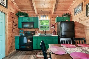Une cuisine de style rustique, avec des meubles de couleur verte, un réfrigérateur noir et un coin repas comprenant une table et des chaises.