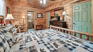 Una habitación tipo cabaña de madera con una cama, una silla, un escritorio, un televisor y una puerta.