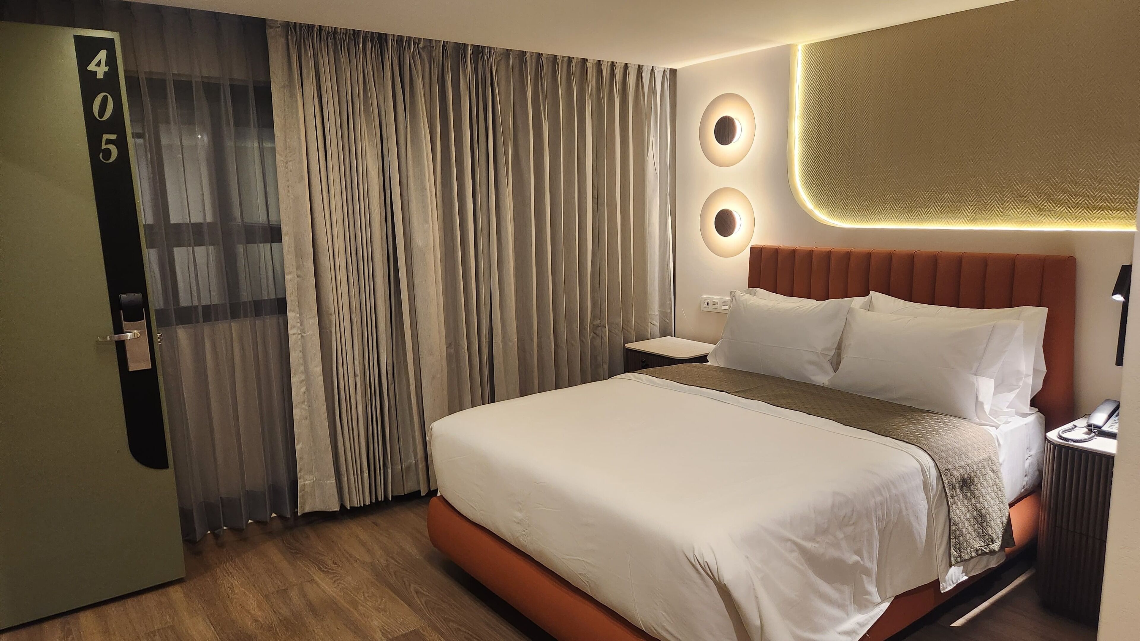 Quarto de hotel com cama grande, mesa de cabeceira, abajur e porta com número.