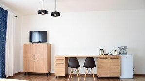 Une pièce moderne et bien éclairée, avec un bureau en bois, deux chaises noires, un téléviseur posé sur un meuble et un réfrigérateur blanc.