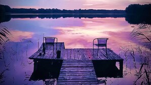 Une jetée en bois s’avançant dans une étendue d’eau calme, avec deux chaises placées face à face, le tout entouré de roseaux.