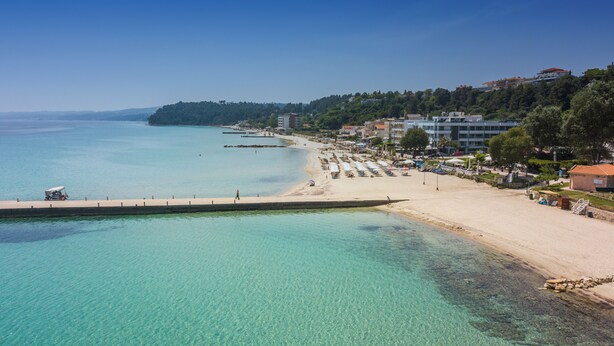 Uma praia costeira com águas turquesa cristalinas, uma faixa de areia e edifícios ao longo da encosta.