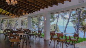 Uma sala de jantar ampla com mesas e cadeiras de madeira, grandes janelas com vista para árvores e água.