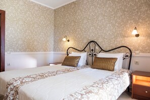 Una cama doble con colcha de estampado floral, dos lámparas de lectura fijadas a la pared y una mesita de noche de madera con cajón.