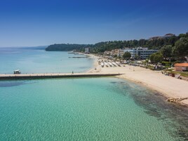 A coastal beach with clear turquoise waters, a sandy shore, and buildings along the hillside.