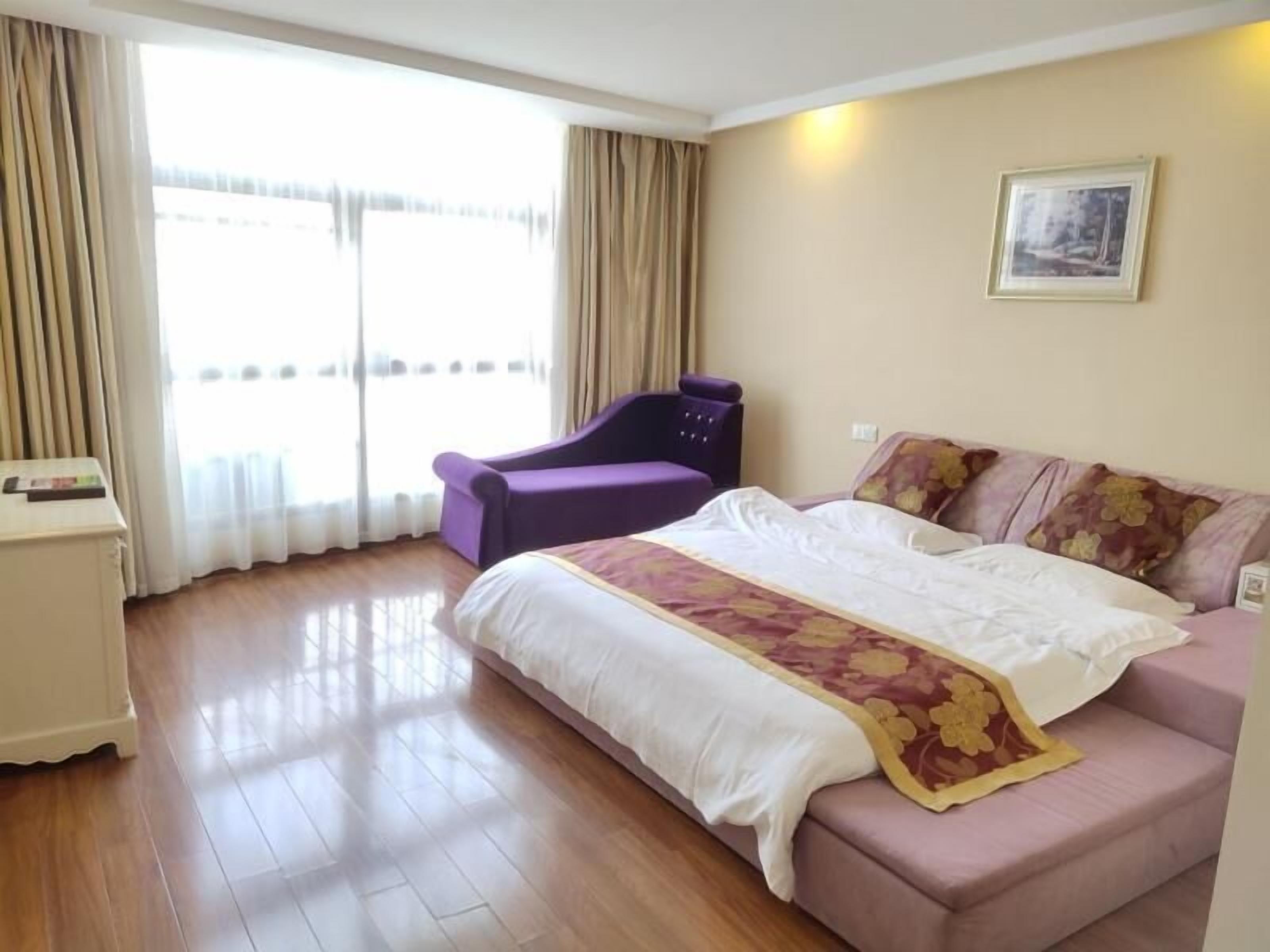 Ein Hotelzimmer mit einem Bett, einem violetten Sessel, einem kleinen Tisch und einem großen Fenster mit Vorhängen.