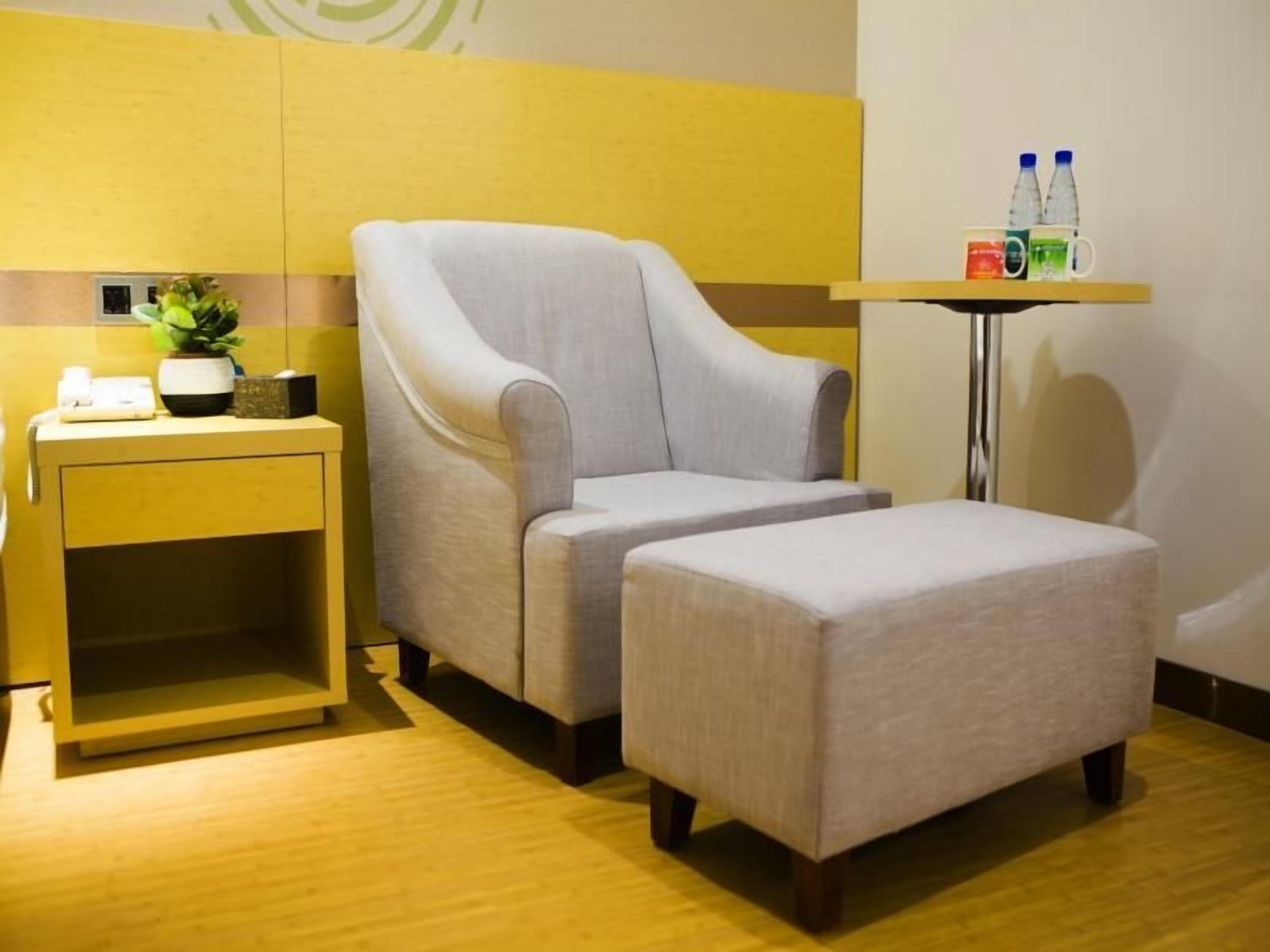 A room with a grey armchair, a matching ottoman, a small side table with a plant, and a wall-mounted shelf with bottles.