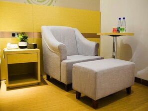 Quarto com poltrona cinza, um banquinho combinado, uma pequena mesa lateral com uma planta e uma prateleira na parede com garrafas.