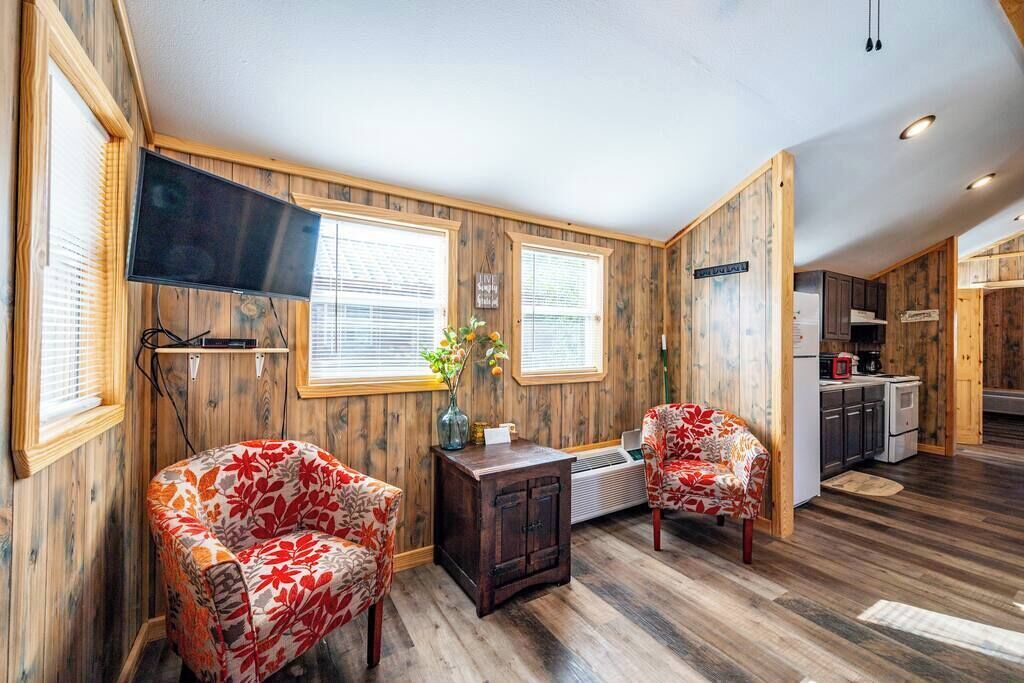 Ein Zimmer mit Holzwänden, einem Wand-Fernseher, zwei stuhlförmigen Sitzgelegenheiten mit Blumenmuster, einer kleinen Holzkommode und einem Fenster mit Jalousien.