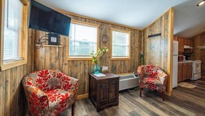 A room with wooden walls, a flat-screen TV, two floral armchairs, a small wooden cabinet, and a window with blinds. - Cedar: Sleeps 3, Tiny Cabin, Pets, Near Park/Lake (Broken Bow)