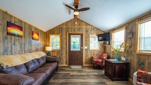 A cozy room with a brown leather sofa, a wooden ceiling fan, a television, and a door with windows. - Cedar: Sleeps 3, Tiny Cabin, Pets, Near Park/Lake (Broken Bow)