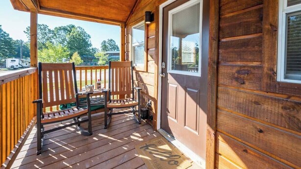 A wooden porch with two chairs and a table, a door with a window, and a view of trees and a vehicle in the background. - Cedar: Sleeps 3, Tiny Cabin, Pets, Near Park/Lake (Broken Bow)