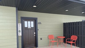 Une porte avec une pancarte « Bienvenue » et une autre indiquant « Il est interdit de fumer », un paillasson et deux chaises rouges devant un bâtiment entouré d’une clôture en bois.