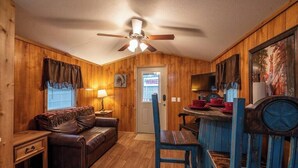 A cozy cabin interior with a wooden interior, a ceiling fan, a dining table with chairs, and a television. - Choctaw: Sleep 4, Tiny Cabin, Pets, Near Park/Lake (Broken Bow)