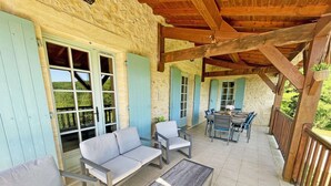 Ein überdachter Patio-Bereich mit Gartenmöbeln, darunter Sofas und ein Tisch, vor einer Steinwand mit blauen Fensterläden.