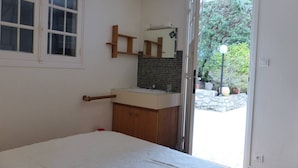 Een slaapkamer met een bed, een houten garderobe, een kleine badkamer met een wastafel en spiegel, en uitzicht op een tuin door een geopende deur.