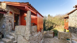 Ein steinernes Gebäude mit einer roten Holztür und einem kleinen Fenster, umgeben von Steinmauern und Stufen.