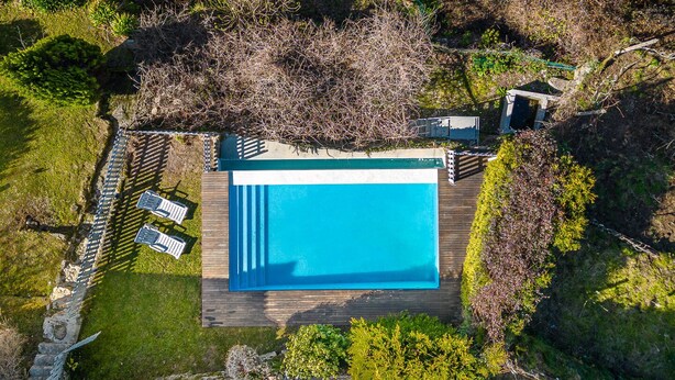 Eine Vogelperspektive auf einen rechteckigen Swimmingpool mit Holzterrasse, umgeben von einer Steinmauer und einem kleinen Garten mit einigen Bäumen und Sträuchern.