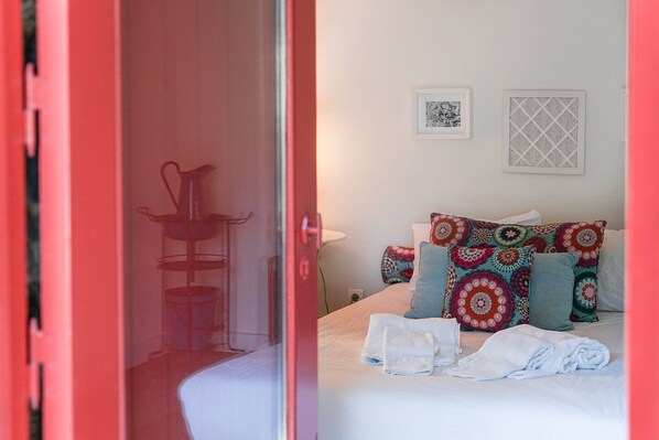 Habitación de hotel con cama, almohadas coloridas, una mesita con un florero y una puerta que da a otra habitación.