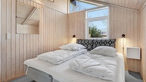 A bedroom with a wooden slat ceiling, a large window, a bed with white bedding, and a bedside table with a lamp.